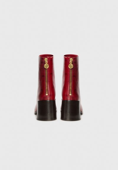 Röda läderankel boots med en glansig finish, med en guldfärgad dragkedja och en staplad svart klack. Enkel design med mjuk textur.