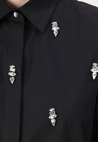 Camicia nera con colletto strutturato, arricchita da quattro dettagli in cristallo trasparente di forme e dimensioni diverse.