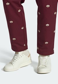 White sneakers made of textured material, featuring a beige accent on the heel. Paired with maroon pants adorned with metallic floral embroidery.