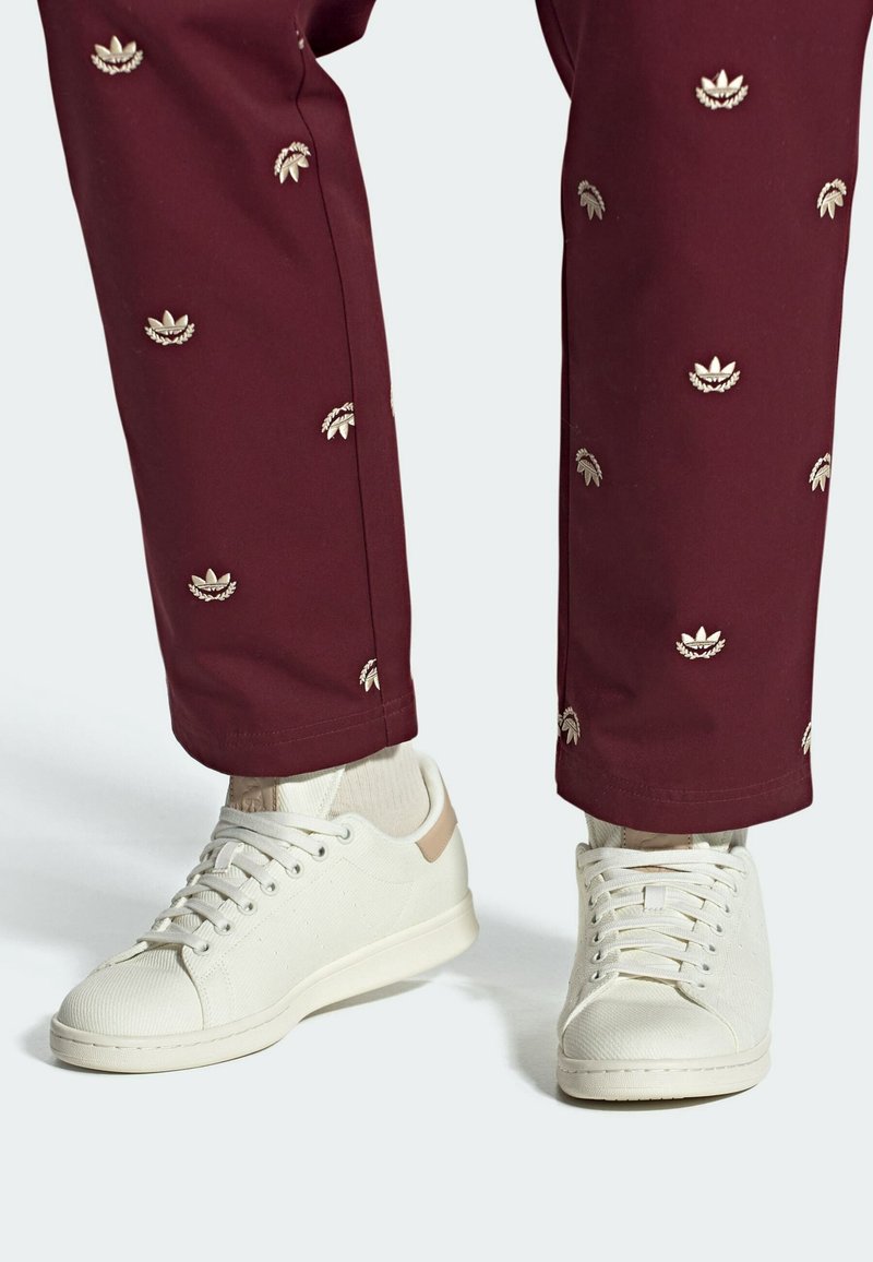 White sneakers made of textured material, featuring a beige accent on the heel. Paired with maroon pants adorned with metallic floral embroidery.