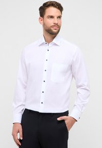 Camicia bianca con bottoni, con una tasca, caratterizzata da bottoni scuri a contrasto e maniche lunghe, abbinata a pantaloni neri.