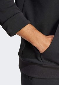 Sudadera negra con un tejido suave, puños acanalados y un bolsillo frontal. El material parece ligero y cómodo.