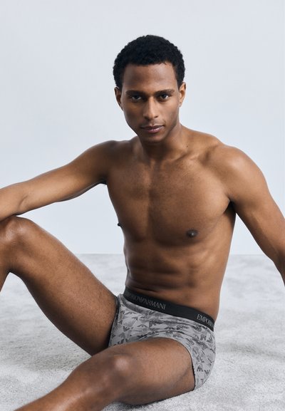 Γκρι κολάν μποξέρ με σχέδιο και μαύρη ζώνη, που διαθέτει λογότυπο. Το μοντέλο κάθεται σε μία μαλακή, ανοιχτού γκρι επιφάνεια με φόντο ουδέτερης απόχρωσης.