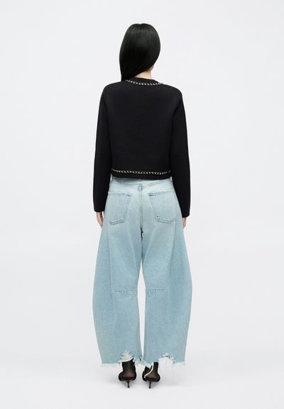 Fekete, rövid fazonú pulóver lánc díszítéssel a nyakkivágásnál, világoskék, széles szárú farmerrel párosítva, amelynek a szegélye rojtos és hátul zsebek találhatók rajta.