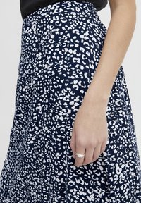 Žena v sukni s navy modro-bílým vzorem, s rukou odpočívající vedle sebe, na prstu má stříbrný prsten.