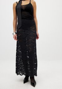 Schwarzer Spitzen-Maxi-Rock mit eng anliegendem Bund und fließender Silhouette. Durchsichtige obere Schicht mit floralen Mustern, kombiniert mit einem schwarzen Satintop und Heels.