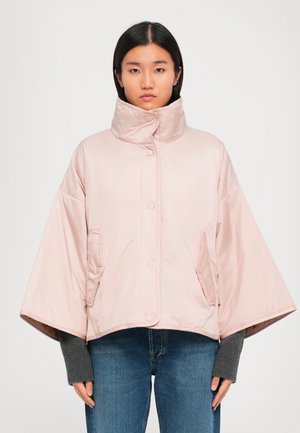 Vrouw in een lichtroze cropped gewatteerde jas met brede mouwen, hoge kraag, drukknoopjes en zijzakken over een grijze lange mouwtrui en jeans.