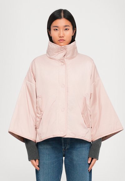 Woman wearing a light pink cropped padded jacket with wide sleeves, high collar, snap buttons, and side pockets over gray long sleeves and jeans.