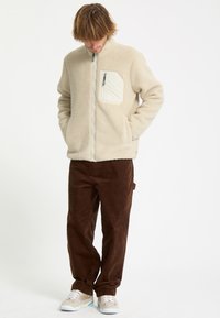 Homme portant une veste polaire beige avec une poche poitrine, pantalon marron et baskets beiges, debout les mains dans les poches, regardant vers le bas.