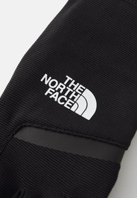 Sort handske med tekstureret stof, der har et hvidt "The North Face"-logo og et udskæringsdetalje for øget fleksibilitet.
