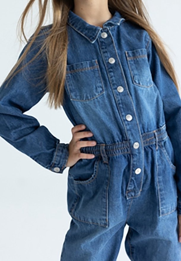 Dítě oblečené v modrém denimovém overal s knoflíky a elastickým pasem, stojící s rukou na boku na jednobarevném pozadí.