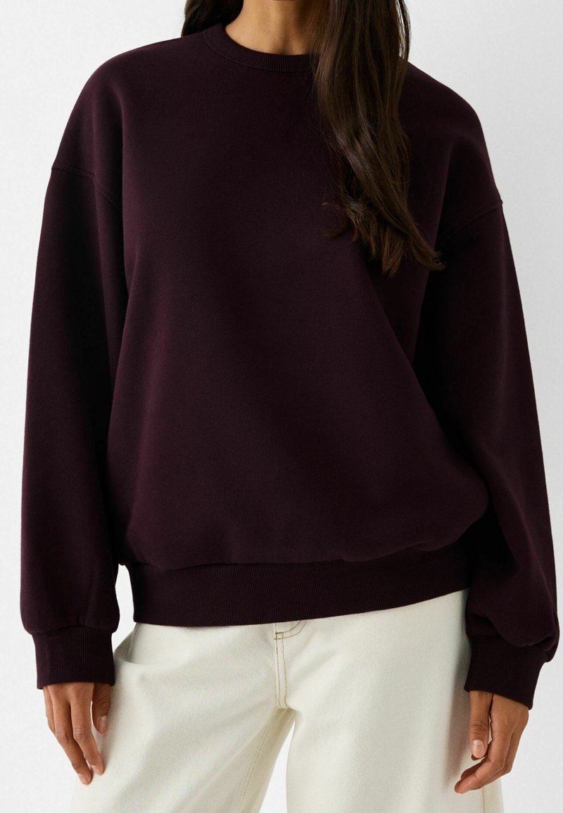 Burgundy oversized sweatshirt med rund hals, ribbstickade ärmslut och nederkant, mjuk textur, i kombination med krämfärgade byxor.