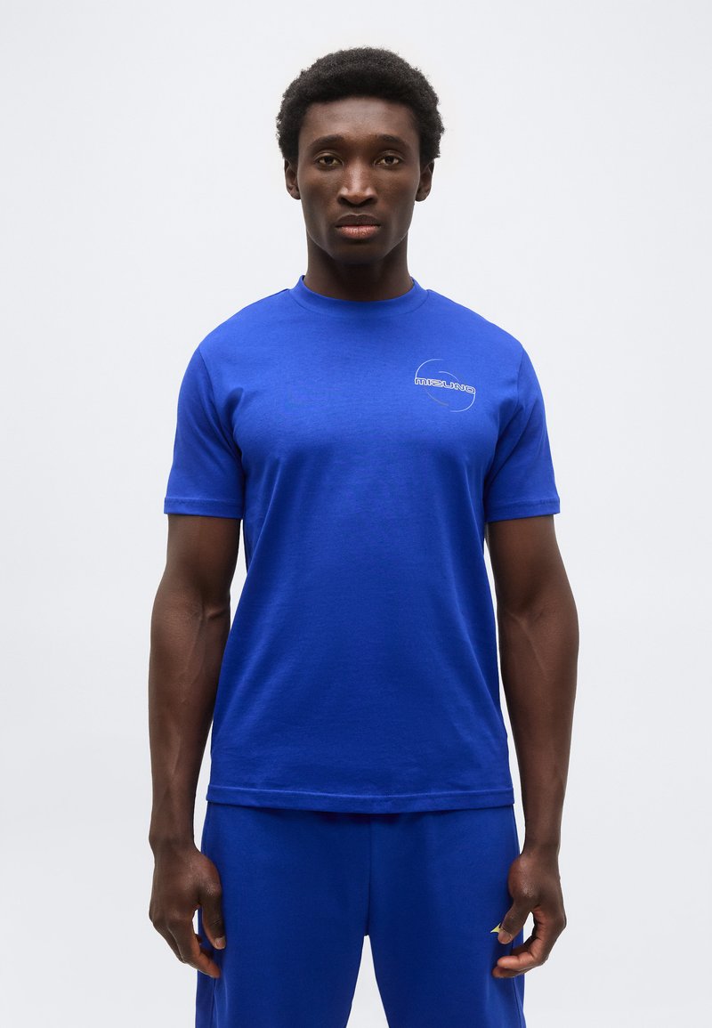 Νεαρός άνδρας με κοντά σγουρά μαλλιά, ντυμένος με μπλε μπλουζάκι Mizuno και αντίστοιχο μπλε παντελόνι, στέκεται μπροστά σε απλό φωτεινό φόντο.