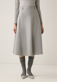 Frau trägt einen hellgrauen A-Linien-Midirock mit Knopfdetails an der Taille, kombiniert mit einem passenden grauen Langarmoberteil, Socken und Schuhen.