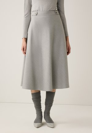 Frau trägt einen hellgrauen A-Linien-Midirock mit Knopfdetails an der Taille, kombiniert mit einem passenden grauen Langarmoberteil, Socken und Schuhen.