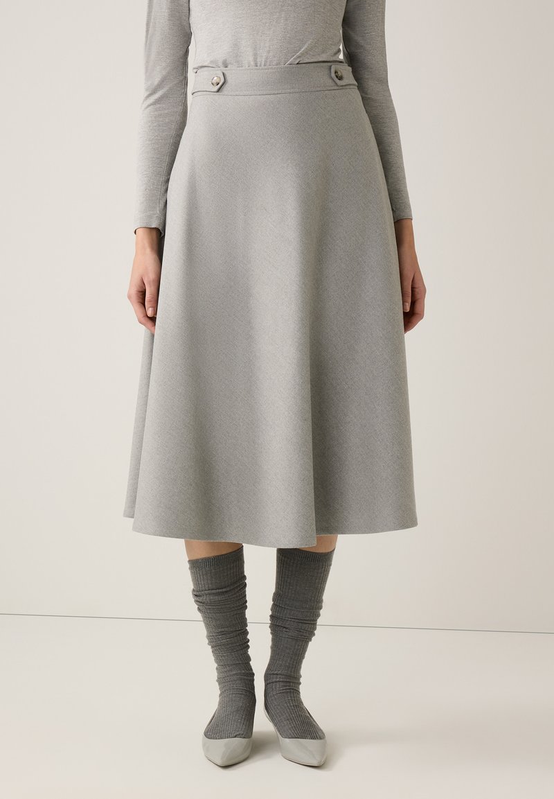 Frau trägt einen hellgrauen A-Linien-Midirock mit Knopfdetails an der Taille, kombiniert mit einem passenden grauen Langarmoberteil, Socken und Schuhen.