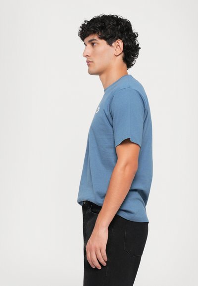 Blaues Baumwoll-T-Shirt mit kurzen Ärmeln, rundem Halsausschnitt und einem kleinen Logo-Detail auf der Brust. Getragen mit schwarzen Hosen.