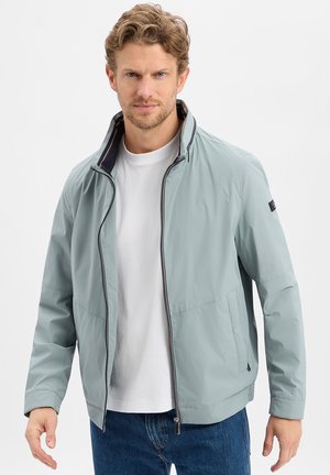 Mężczyzna w jasnoniebieskiej kurtce na suwak na białej koszulce i niebieskich dżinsach, stojący na tle jednolitej białej ściany.