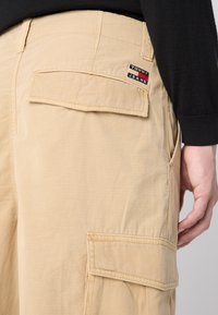 Béžové cargo nohavice vyrobené z ľahkého materiálu, s bočnou vreckom, prešitými akcentmi a nášivkou "Tommy Jeans".