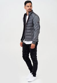 Gilet zippé gris matelassé avec des manches en tricot gris foncé, jeans noirs déchirés et baskets blanches. Comprend des poches latérales et un accent de logo.