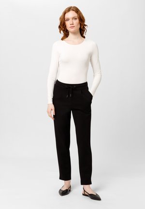 Witte langemouwen shirt in combinatie met zwarte, taps toelopende broek met een verstelbare trekband in de taille en zijzakken; model staat in zwarte ballerina's.