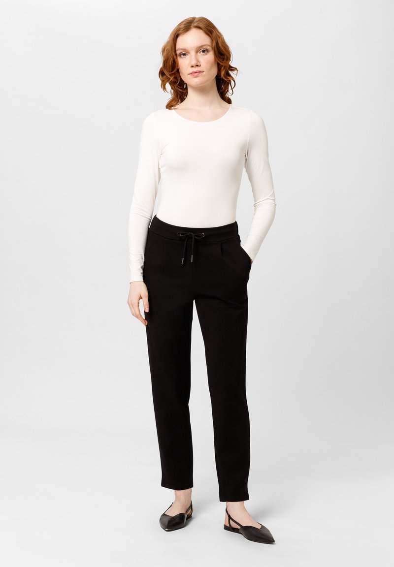 Witte langemouwen shirt in combinatie met zwarte, taps toelopende broek met een verstelbare trekband in de taille en zijzakken; model staat in zwarte ballerina's.