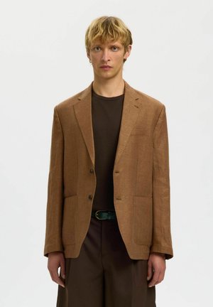 Jeune homme aux cheveux blonds portant un blazer marron sur une chemise marron foncé et un pantalon marron assorti, sur un fond uni.