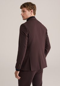 Joven con una chaqueta de traje marrón oscuro y pantalones a juego, girado ligeramente hacia un lado, frente a un fondo claro y sencillo.