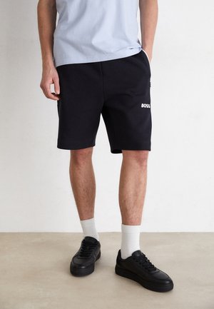 Shorts noirs avec taille élastique, poches latérales et logo "BOSS" blanc. Associés à des baskets noires et des chaussettes blanches mi-hautes. Tenue décontractée.