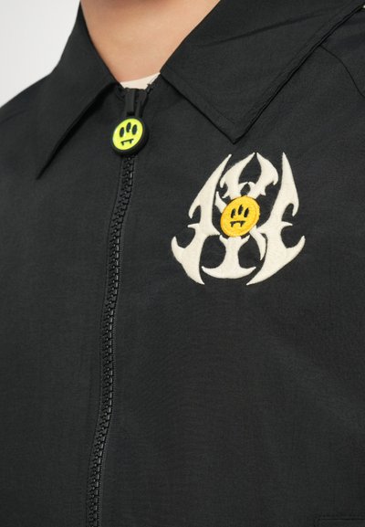 Giacca nera con colletto, dotata di una zip frontale. Elementi decorativi notevoli includono un design ricamato bianco e un bottone giallo a forma di sorriso.