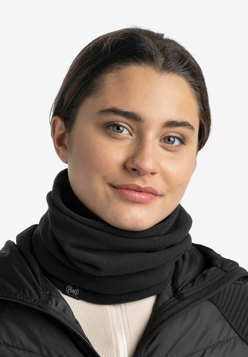 Calentador de cuello de lana negra con una textura suave, que presenta un acento de marca. Se lleva con una chaqueta negra y una parte superior de color claro.