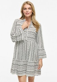 Robe à manches longues avec un motif géométrique noir et blanc, décolleté en V, manches évasées et jupe à volants. Fabriquée en tissu léger.