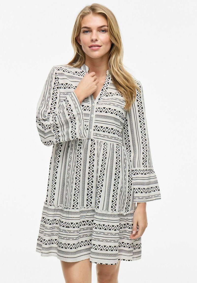 Robe à manches longues avec un motif géométrique noir et blanc, décolleté en V, manches évasées et jupe à volants. Fabriquée en tissu léger.