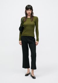 Μπλούζα σε ελιά πράσινο με μακριά μανίκια και ριμπ υφή, συνδυασμένη με μαύρο ψηλόμεσο παντελόνι cropped και μαύρα μπαλέτα. Το μοντέλο κρατά μια μικρή μαύρη τσάντα.