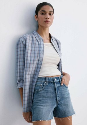 Vrouw staat tegen een witte muur, draagt een blauw geruit overhemd over een wit tanktop en hoog getailleerde spijkershorts met rafelige zomen.