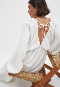 Weißes, strukturiertes Kleid mit Puffärmeln und tiefem Rückenausschnitt, mit einer Schleife gebunden. Modell sitzt auf einem geflochtenen Holzstuhl.