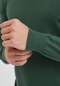 Pull en tricot vert avec des poignets côtelés, montrant une main ajustant la manche. Le tissu semble lisse avec un léger éclat.