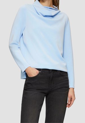 Kobieta w jasnoniebieskiej bluzce z długim rękawem i drapowanym kołnierzem oraz czarnych dżinsach, z jedną ręką w kieszeni, stojąca na jednolitym tle.