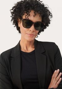 Occhiali da sole con una tonalità verde scuro e montatura tartarugata. Indossati da una persona in un blazer nero e una maglietta, su uno sfondo neutro.