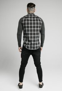 Camisa de manga larga con un reverso de cuadros grises y mangas gris oscuro, combinada con pantalones ajustados negros y zapatos negros. Textura suave y diseño entallado.