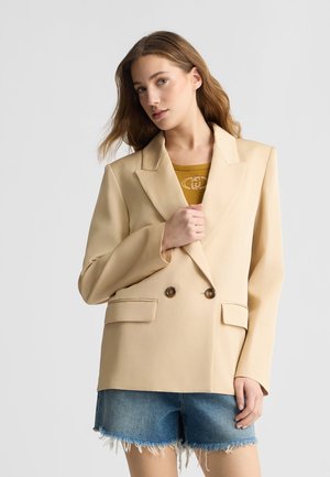 Blazer beige su misura con design doppiopetto, due tasche anteriori e bottoni marroni, indossato sopra un top giallo e pantaloni shorts di denim.