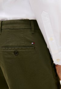 Pantalones de tejido en verde oliva con un bolsillo trasero, botón negro y un pequeño detalle de logo en rojo, blanco y azul cerca del bolsillo.