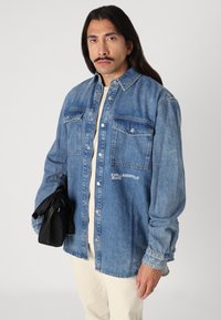 Világoskék farmer gombos ing, két mellzsebbel. A "KARLLAGERFELD JEANS" felirat hímzéssel készült. A modellen egy fekete dzseki van.