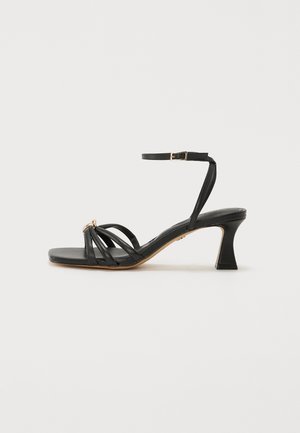 Sandale à talon haut noire en cuir. Présente une ouverture à l'avant, un design avec plusieurs sangles, une boucle à la cheville et un talon carré.