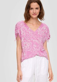 Rosa blusend mit Blumenmuster, V-Ausschnitt und kurzen Ärmeln mit gebundenen Akzenten. Aus leichtem Stoff gefertigt und kombiniert mit weißen Hosen.