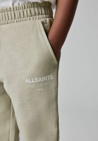 Detailný záber na osobu v béžových teplákových nohaviciach s elastickým pásom, ruka v vrecku, a bielym logom ALLSAINTS s japonským textom.