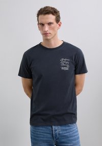 Camiseta de algodón gris oscuro de manga corta con cuello redondo y texto impreso en blanco en el pecho. Corte casual sin patrones visibles.