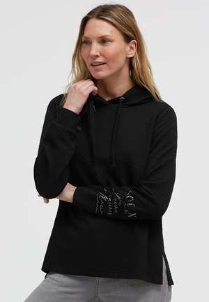 Frau mit blondem Haar, die einen schwarzen texturierten Hoodie mit Kordeln und besticktem Text am linken Ärmel trägt, kombiniert mit hellgrauen Hosen.