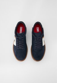 Tamsiai mėlyni zomšiniai sportbačiai su balta akcentine juostele, apvaliu batu pirštu ir guminiais padais. Yra tamsios raištelės ir raudonas padas su "CAMPER" ženklu.