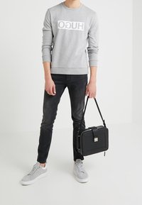 Grauer Sweatshirt mit weißem Logo, schwarze Jeans und graue Sneakers. Hält eine schwarze quadratische Tasche mit einem Riemen und einem metallischen Verschluss.